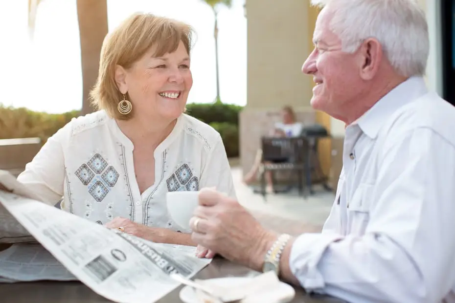 older couple smiling to each other while drinking coffee and reading newspaper