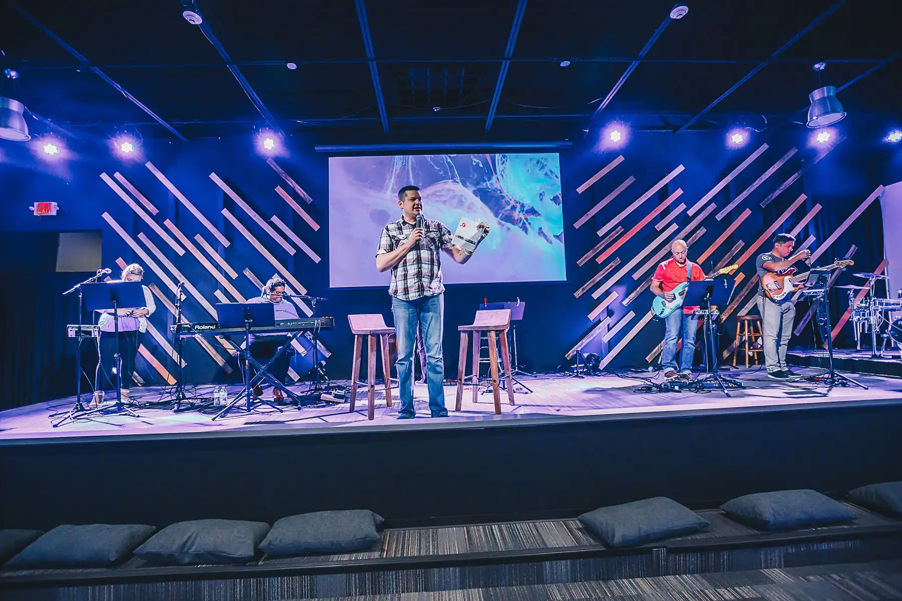 pastor speaking on the stage with people at the back holding instruments